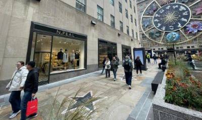 Street view of the N.Peal store at Rockefeller Centre, with pedestrians walking past, a large decorative clock on the building, and landscaped planters along the walkway