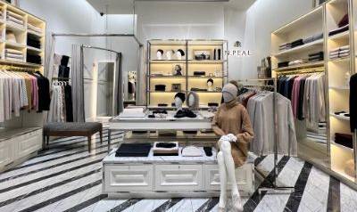 Interior of the N.Peal Rockefeller store with neatly displayed clothing on illuminated shelves and racks, a central table showcasing folded garments, and a seated mannequin dressed in neutral-toned knitwear