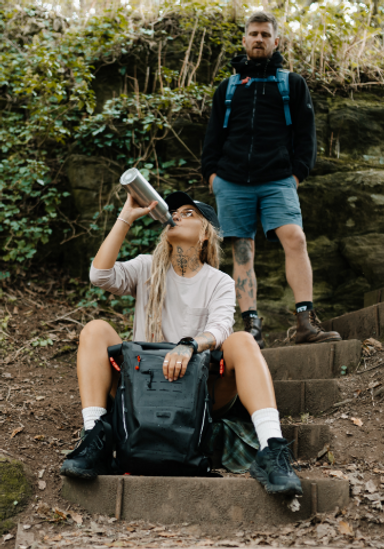 Woman drinking from Red Equipment Stainless Steel Water Bottle whilst sat with Adventure Waterproof Backpack in Obsidian Black. Man carrying the Red Equipment Adventure Waterproof Backpack in Storm Blue.