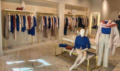 Munich store interior with gold clothing racks displaying neatly arranged garments, a mannequin seated on a bench dressed in a blue top and white trousers, and another mannequin wearing a layered knitwear ensemble